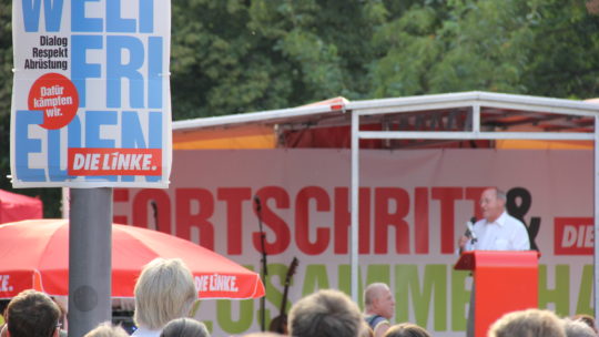 Bild von einer Wahlkampftveranstaltung mit Gregor Gysi, im Vordergrund zu sehen ein Plakat zur Landtagswahl 2019 mit der Aufschrift "Weltfrieden"